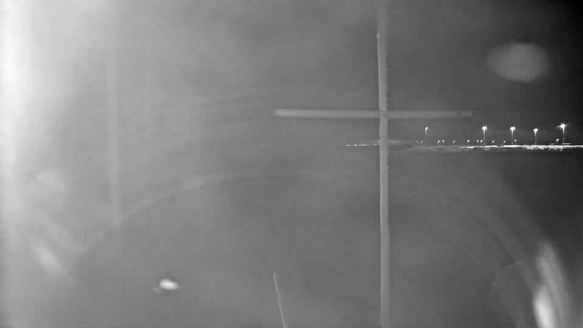 An overcast dusk or dawn scene reveals a sandy beach meeting a calm sea with distant port lights under a cloudy sky, partially framed by a blue foreground structure and a grey cross-shaped pole.
