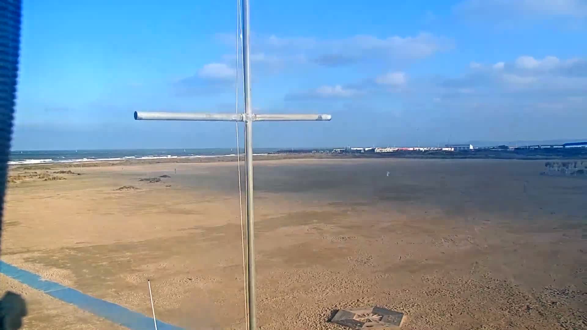 An overcast dusk or dawn scene reveals a sandy beach meeting a calm sea with distant port lights under a cloudy sky, partially framed by a blue foreground structure and a grey cross-shaped pole.