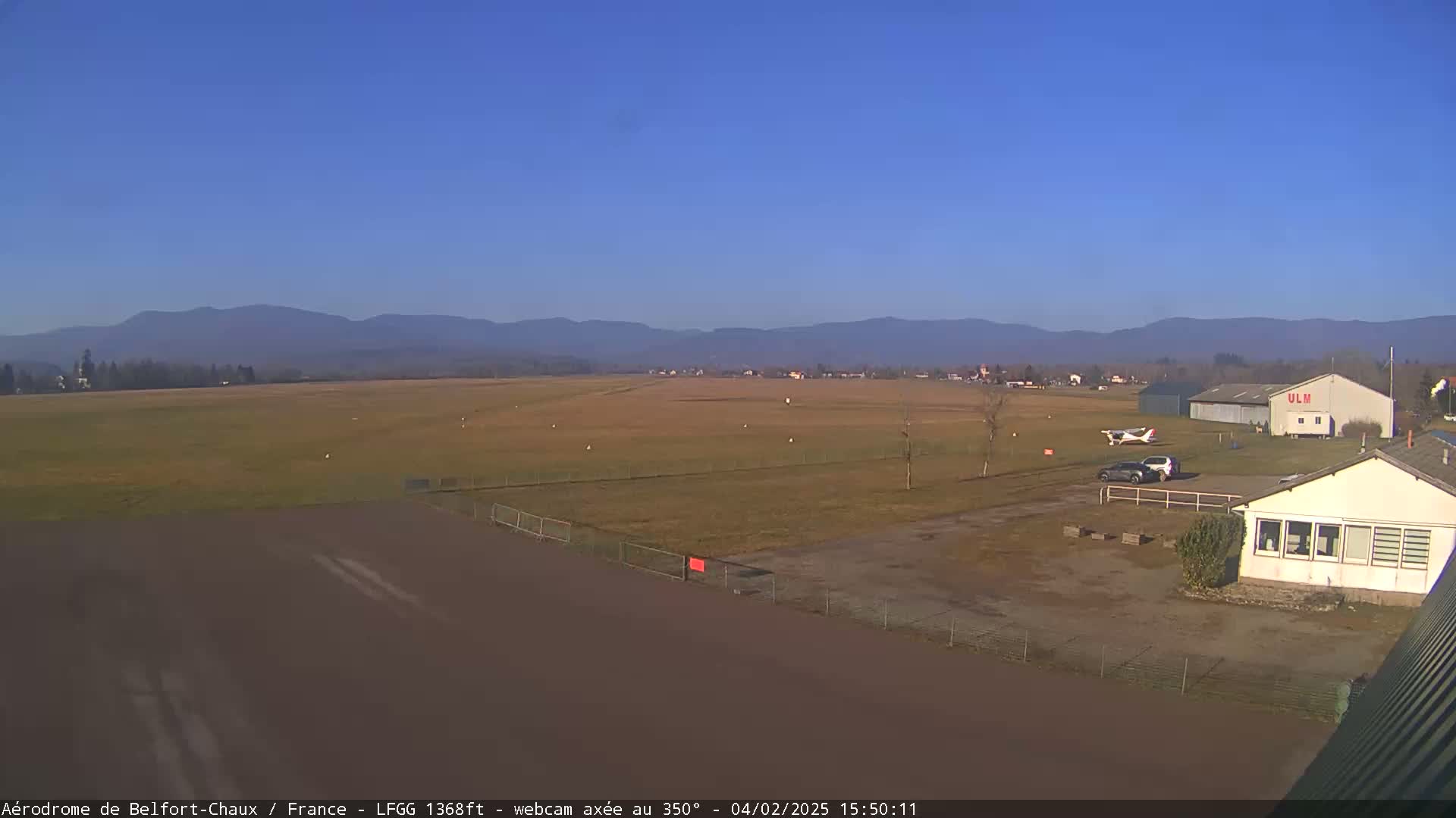 A small airfield with a single-engine airplane parked near a hangar, under a clear blue sky and in front of distant mountains.