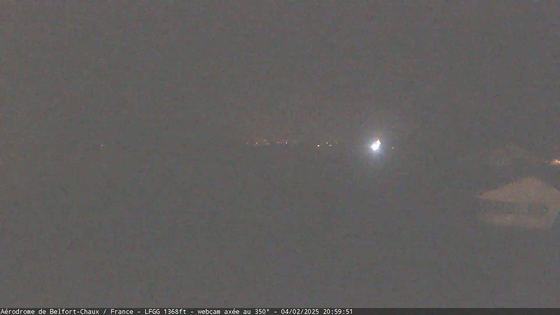 A nighttime, low-light outdoor image shows a distant line of lights and a brighter light to the right, possibly a vehicle headlight, against a dark, overcast sky.