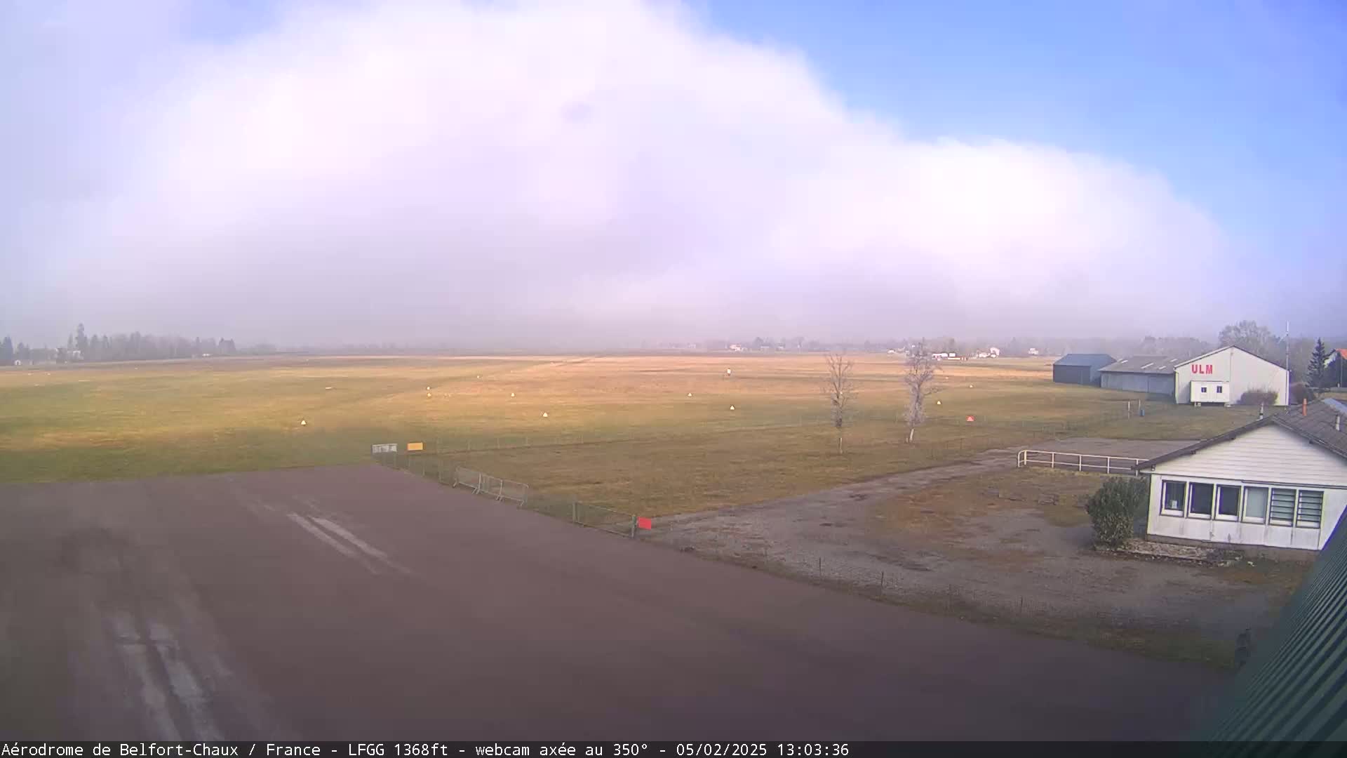 A hazy airfield with a runway, grassy fields, and several buildings is visible under a partly cloudy sky.