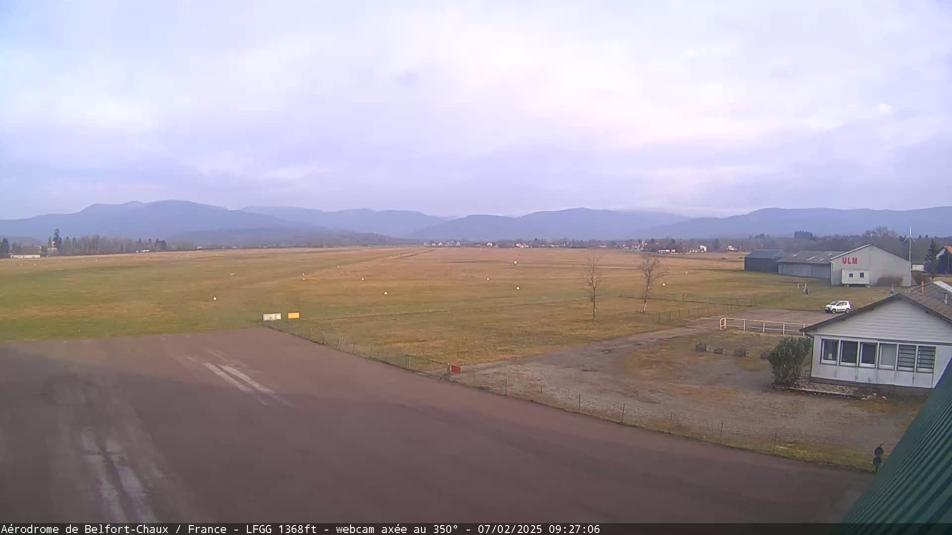 A flat, grassy airfield with a runway and buildings is situated in front of a range of distant mountains under a mostly clear sky.