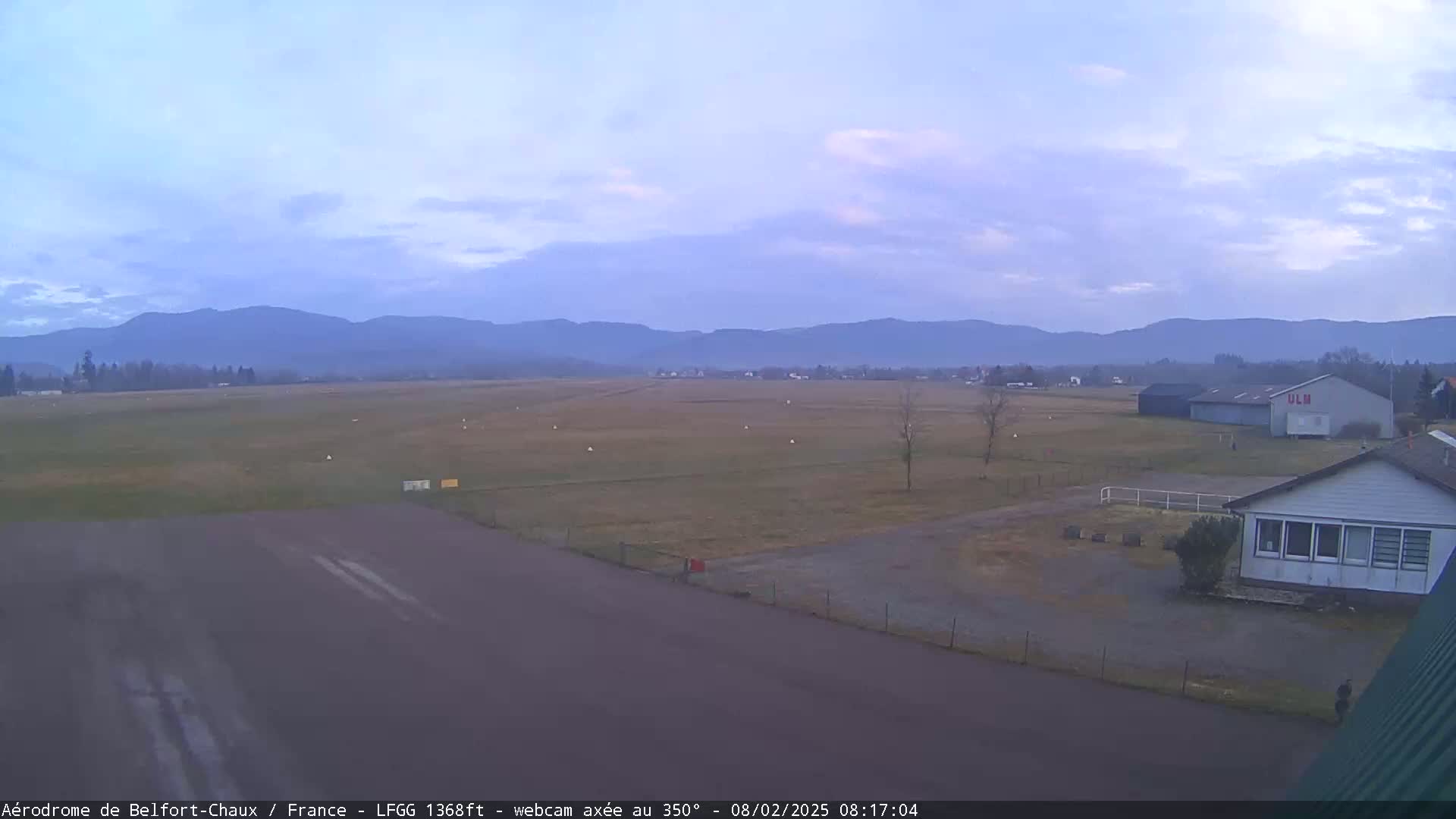 An airfield with a runway, buildings, and a few scattered trees stretches out before a range of distant, hazy mountains under a mostly clear, pale sky.