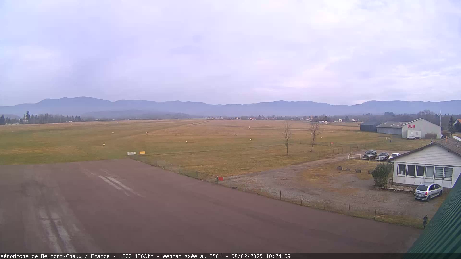 A mostly clear day reveals a flat airfield with a runway and several buildings, set against a backdrop of distant, hazy mountains.
