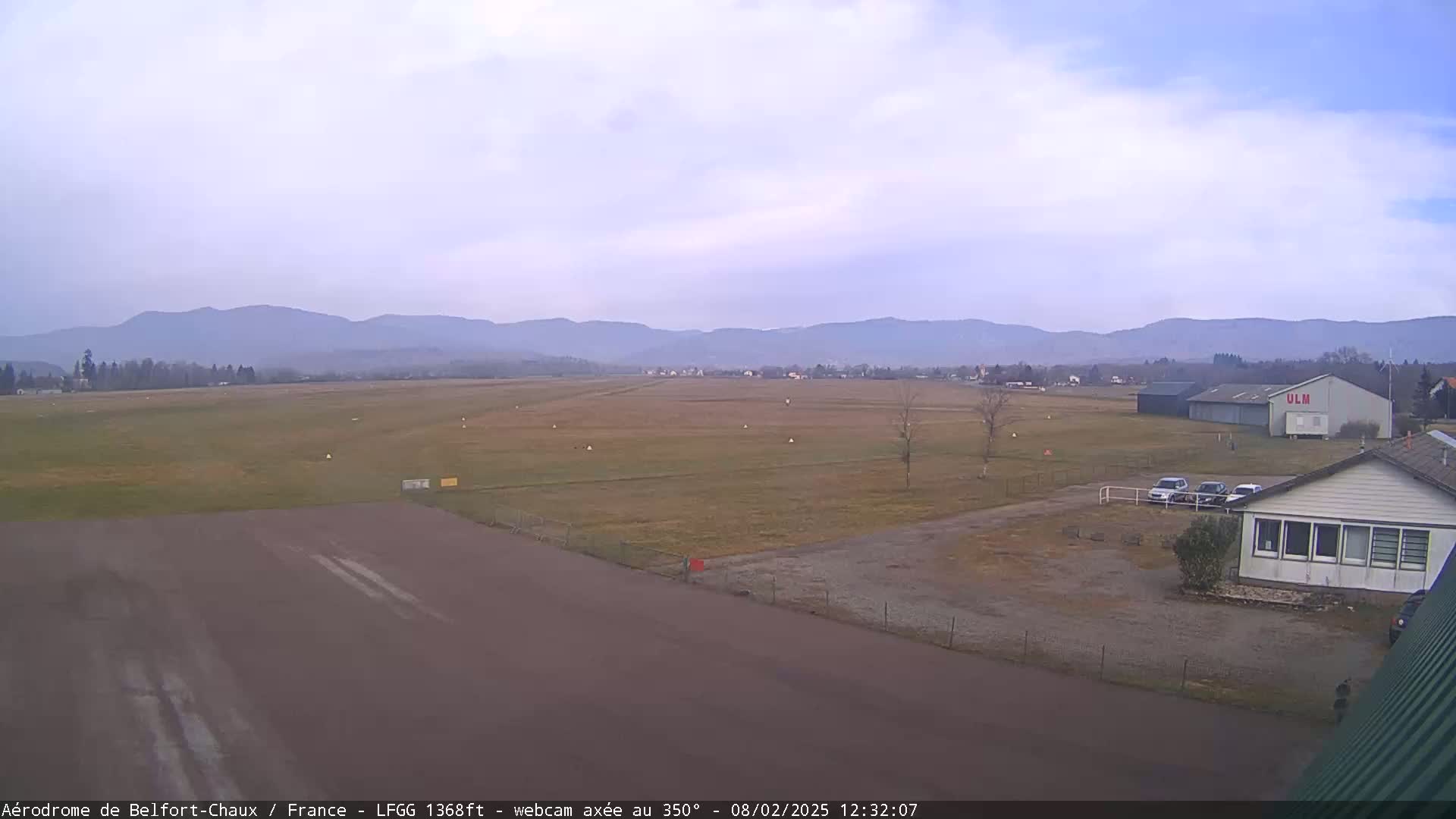 A mostly clear, overcast day reveals a flat, grassy airfield with a runway, a hangar, and several small buildings, all set against a backdrop of distant mountains.