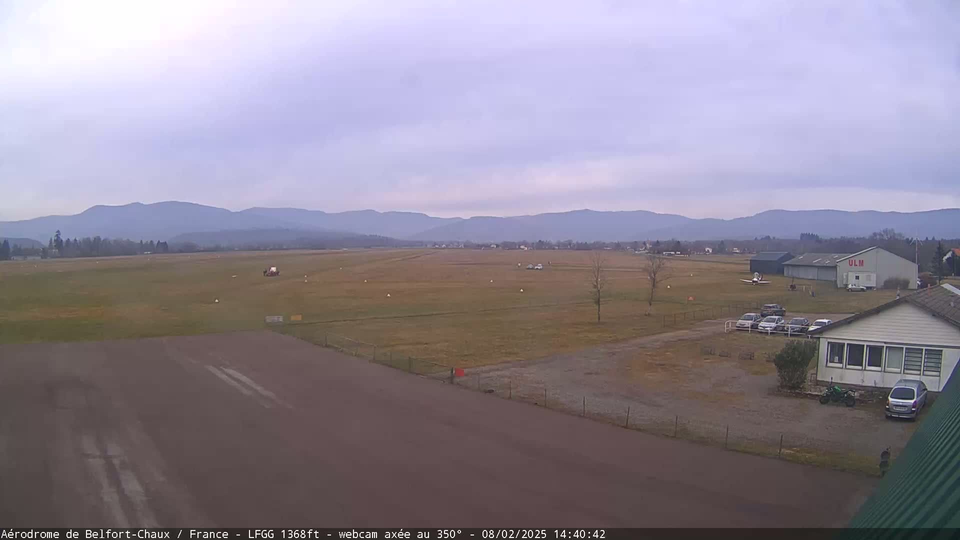 A small airfield with a runway, a hangar, and several parked cars is visible under an overcast sky with distant mountains in the background.