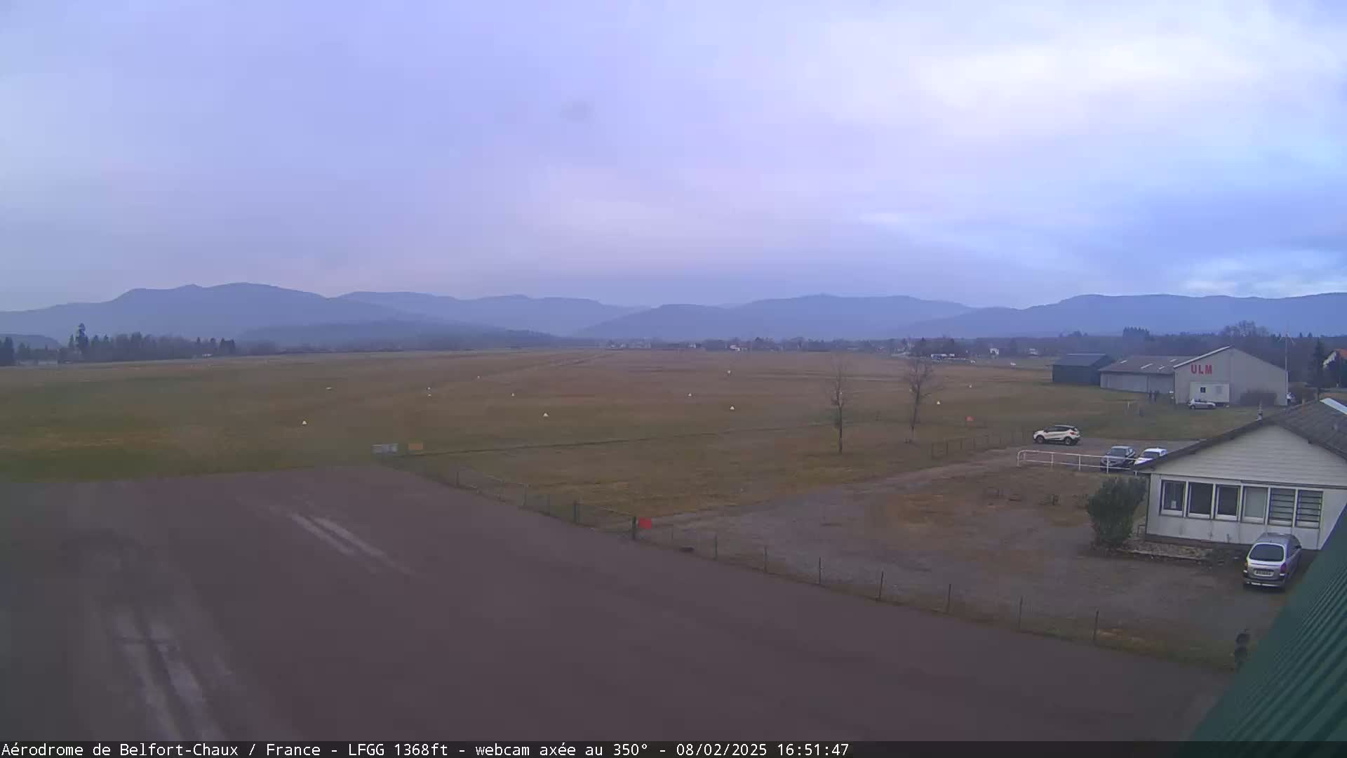 A mostly clear, overcast sky over a flat airfield with a few buildings and cars near a runway, backed by distant, hazy mountains.