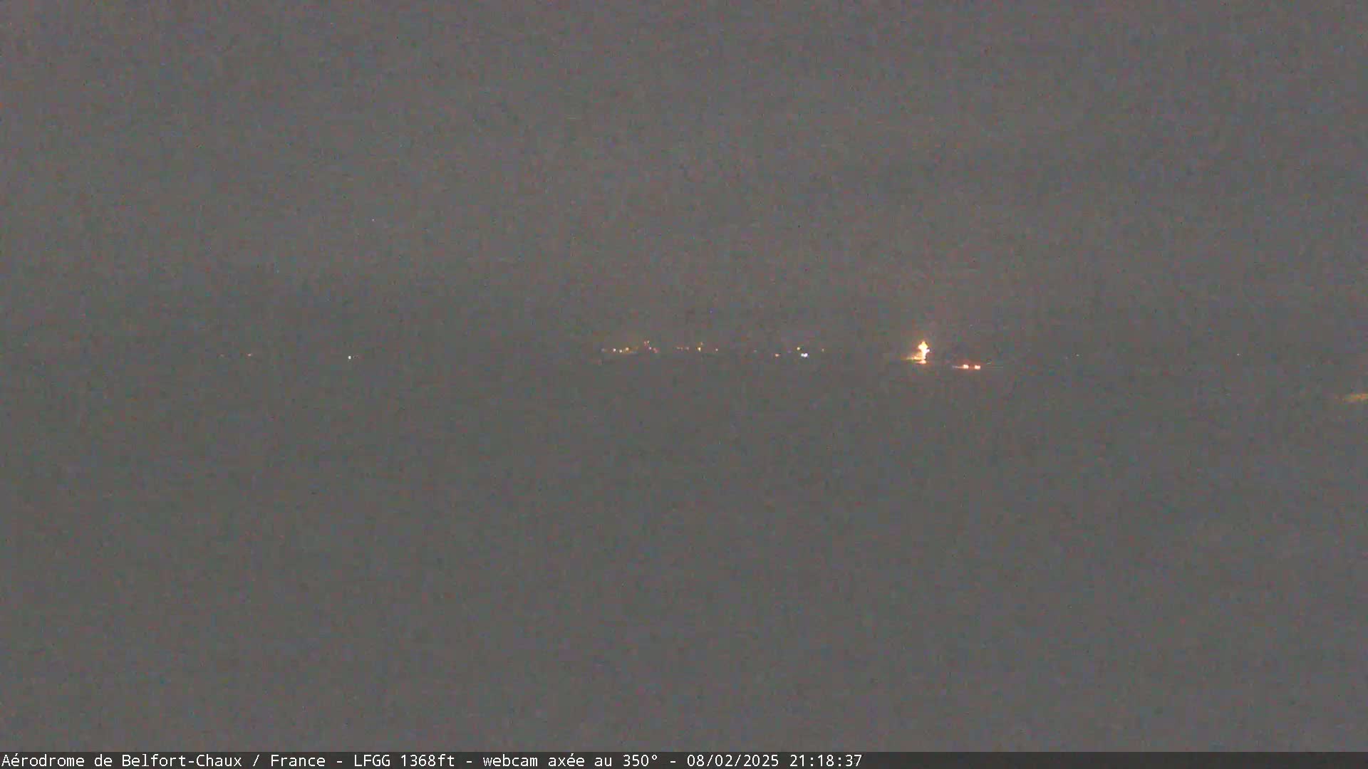 A nighttime, low-light view shows a distant line of faint lights, possibly a town or village, under a dark, overcast sky.