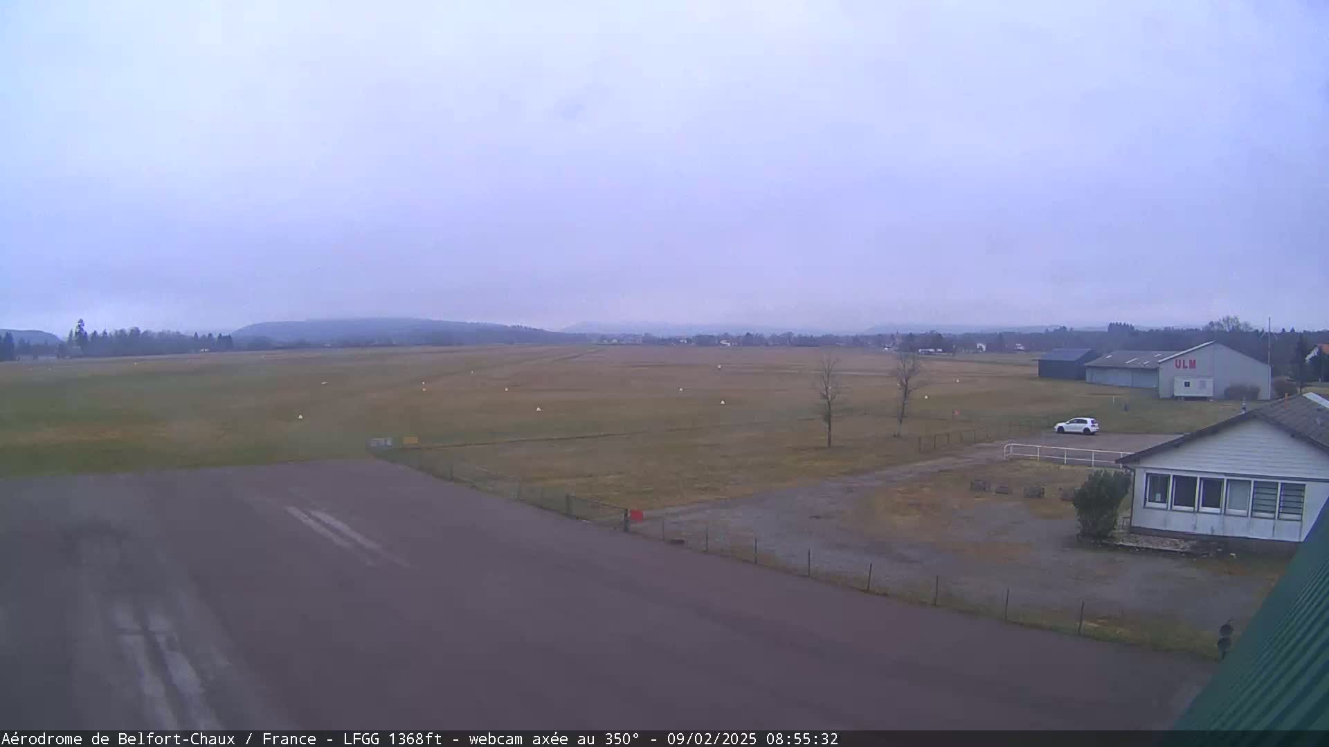 A mostly clear, overcast day reveals a flat airfield with a runway, a few buildings, and a single car parked nearby.
