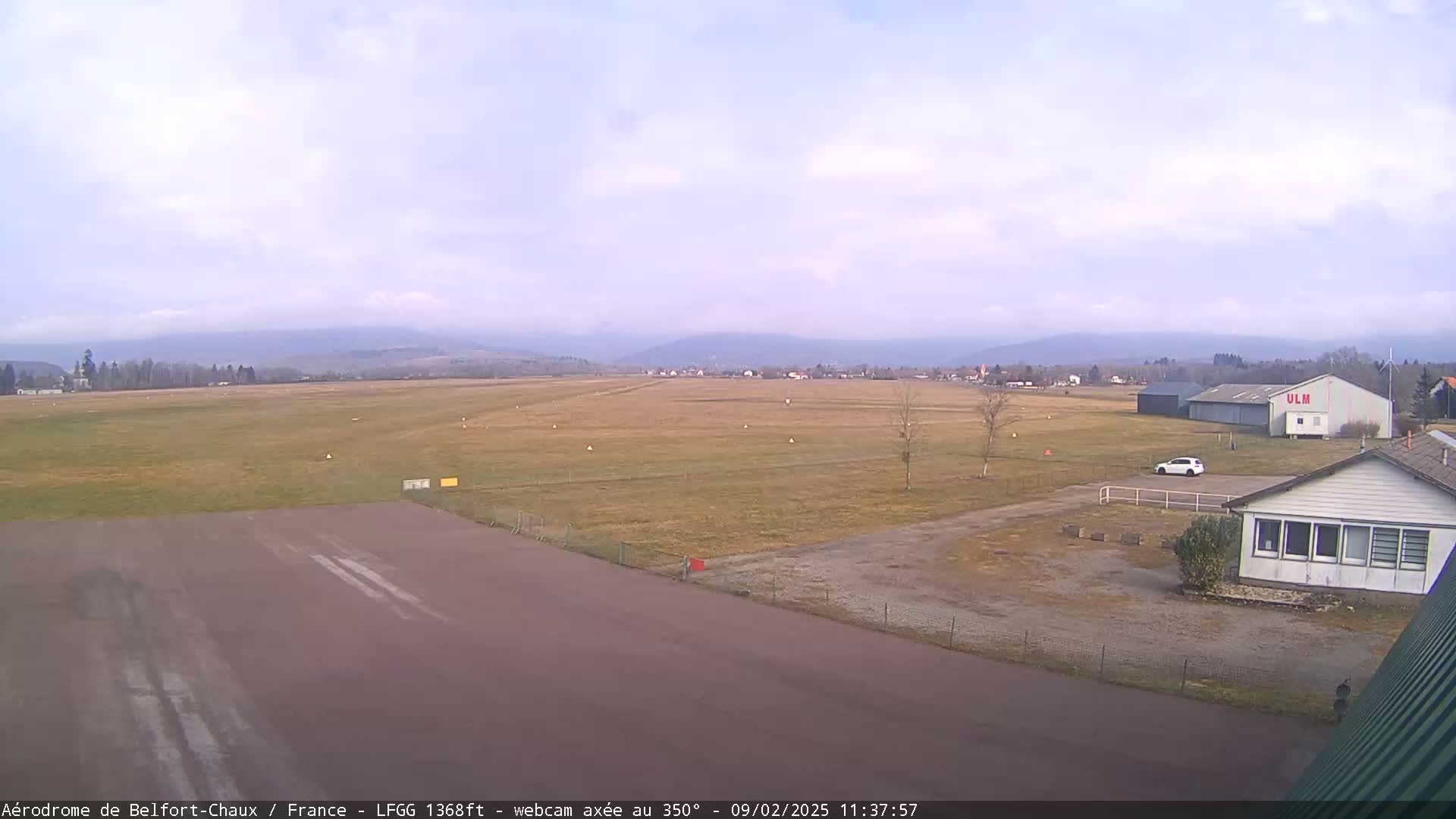 A mostly clear day reveals a runway and airfield, with a small building and a single car visible, set against a backdrop of distant mountains.