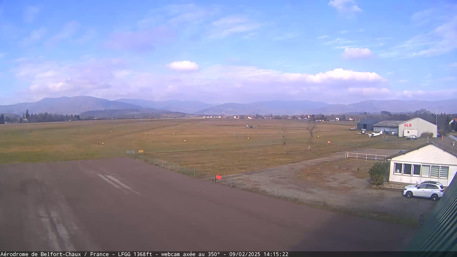 A small airfield with a few light aircraft and a hangar is situated in a valley under a partly cloudy sky, with distant mountains visible in the background.