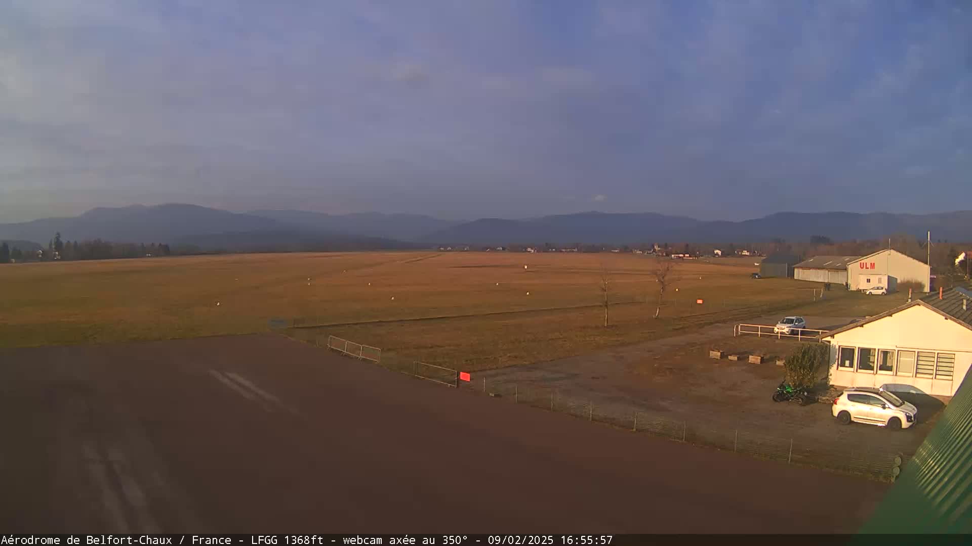 A small airfield with a few cars and buildings is situated in front of a flat, grassy field, with distant mountains under a clear, light-blue sky.