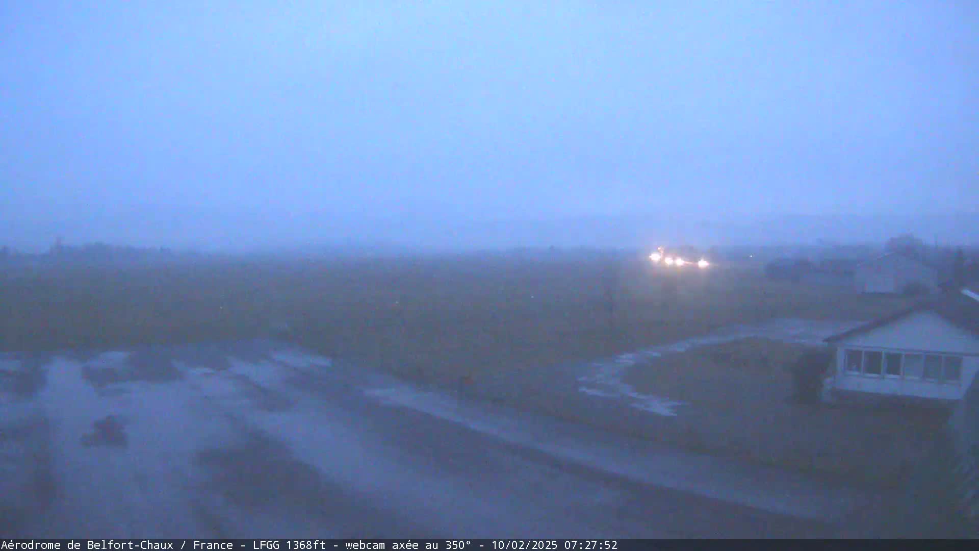 A foggy, overcast scene shows a wet road and houses in the distance, with a few headlights visible through the mist.