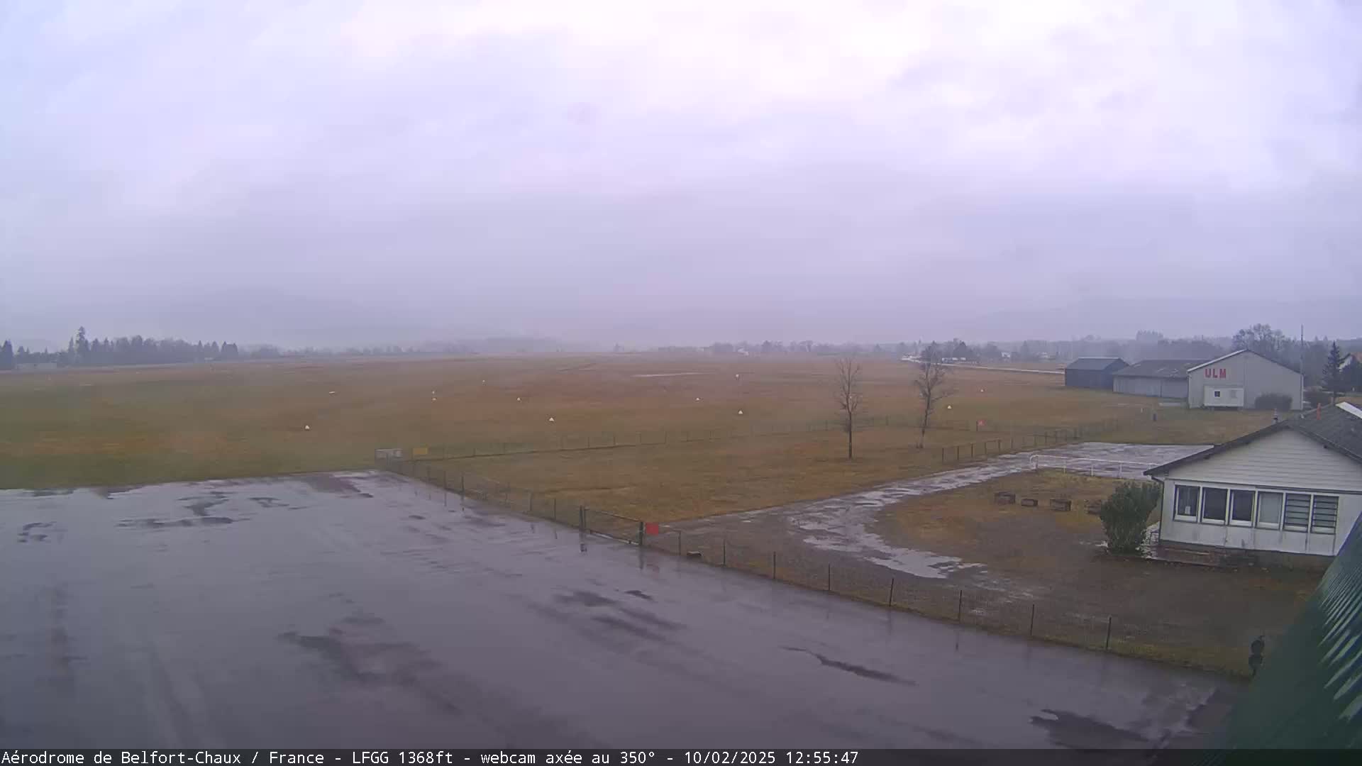 A wet airfield is visible under an overcast sky, with several buildings and a grassy field in view.