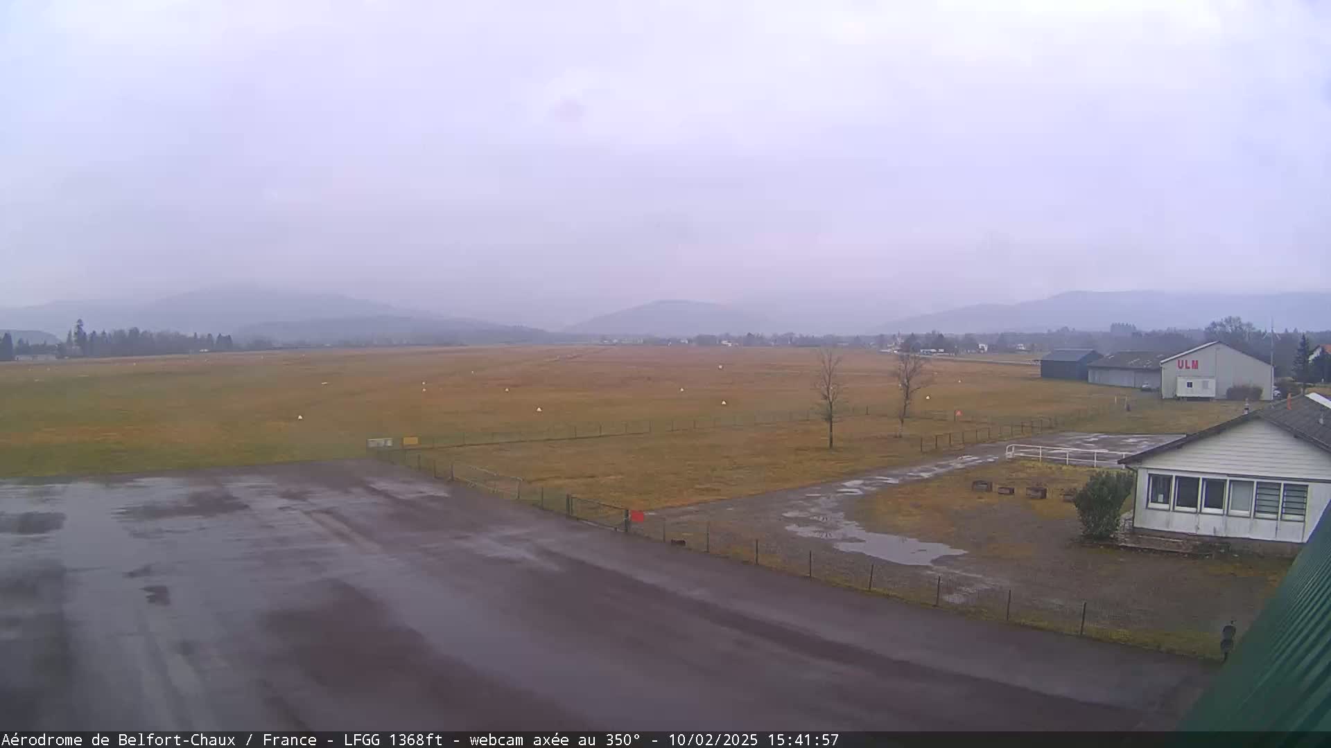 A wet airfield with a small building and distant hazy mountains under an overcast sky.