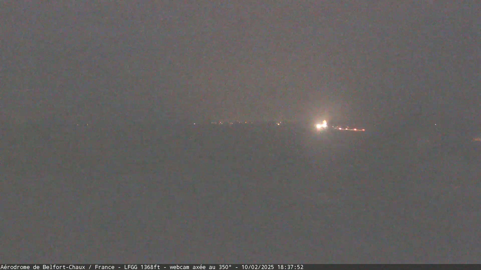 A dark, hazy night scene shows a distant line of faint lights, possibly a town or airport runway, with a brighter light source to the right.