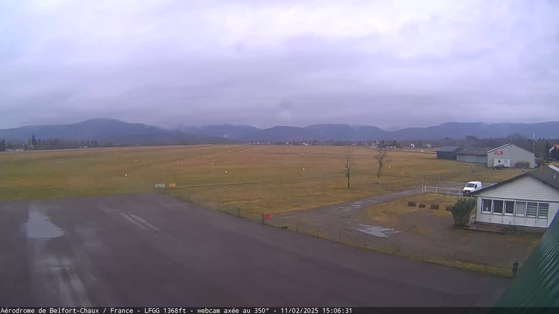 A long airfield with a few buildings and vehicles is visible under an overcast sky, with mountains in the background.