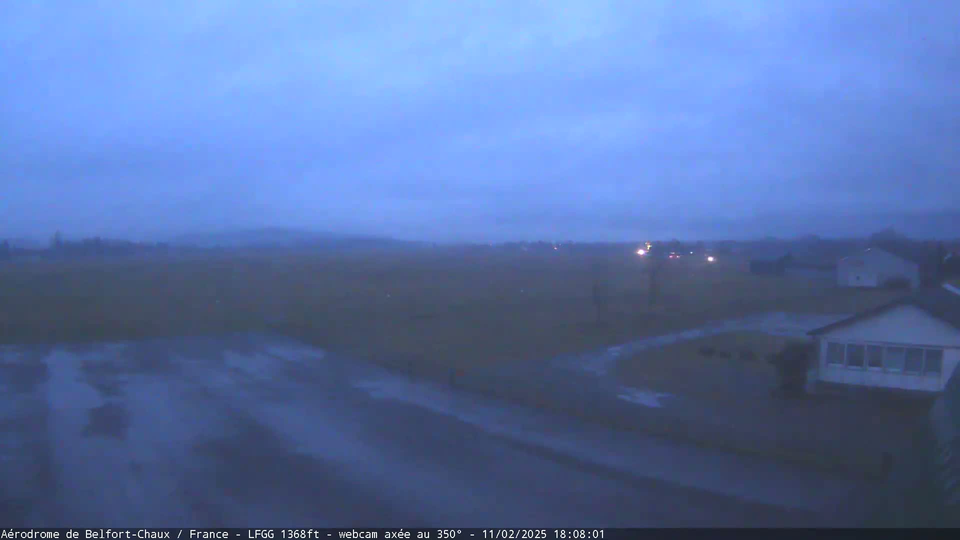 A wet, dark scene shows a flat expanse of land with a few houses and some distant lights under a gloomy sky.