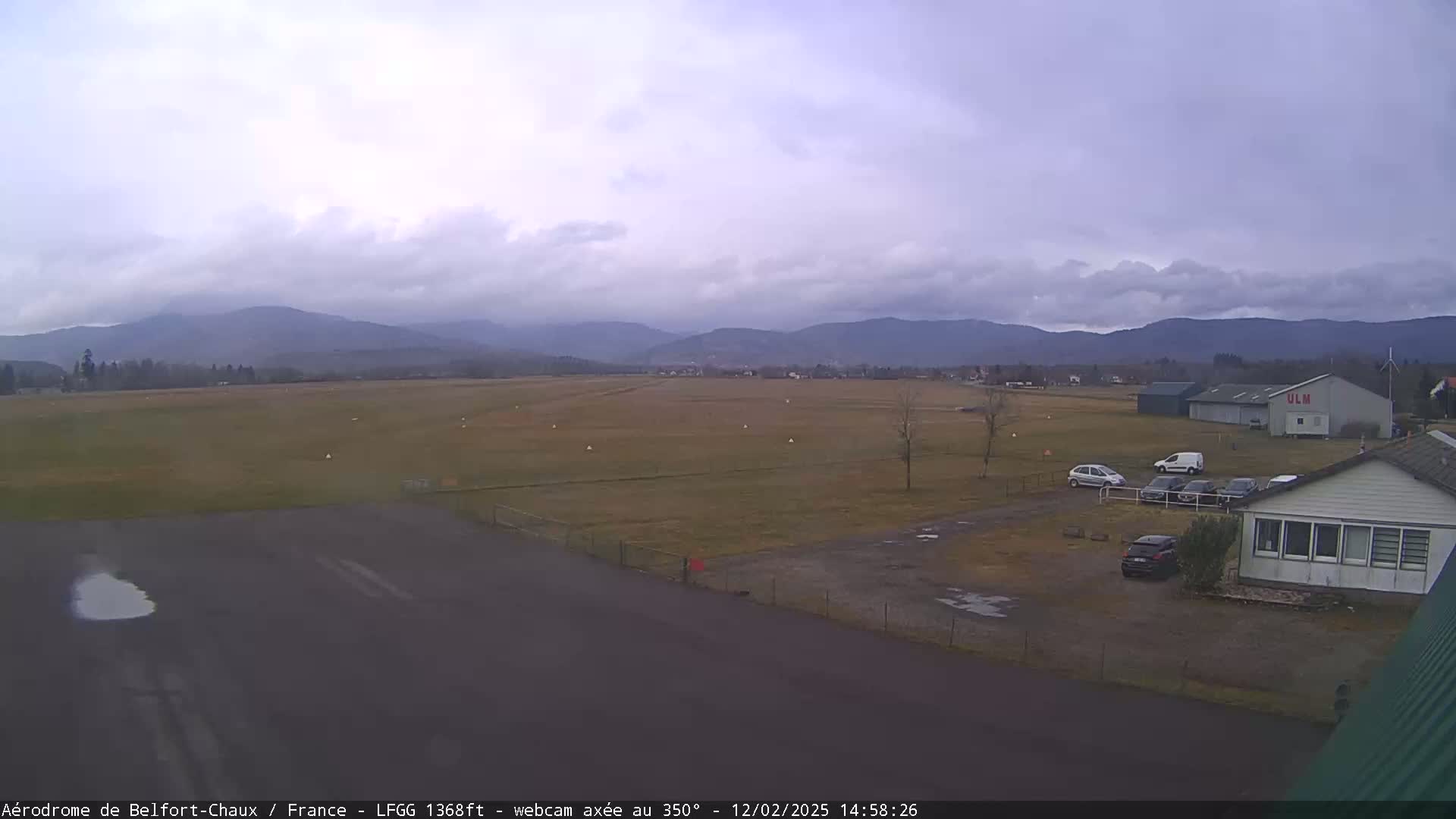 A flat, grassy airfield with a few cars parked near a building is visible under an overcast sky, with mountains in the background.