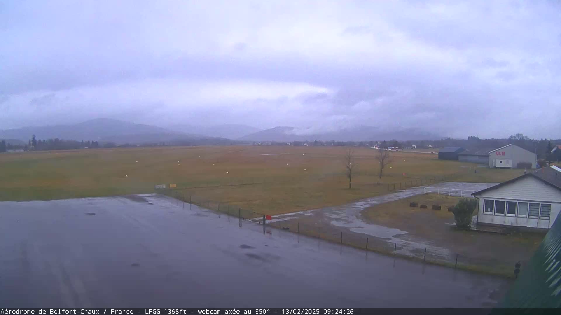 A wet airfield with a small building and distant mountains under an overcast sky.