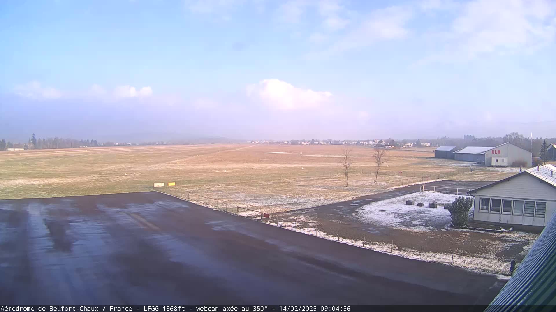 A mostly clear, slightly hazy day with patches of snow on the ground at a small airfield, showing a runway, a hangar, and a small building.
