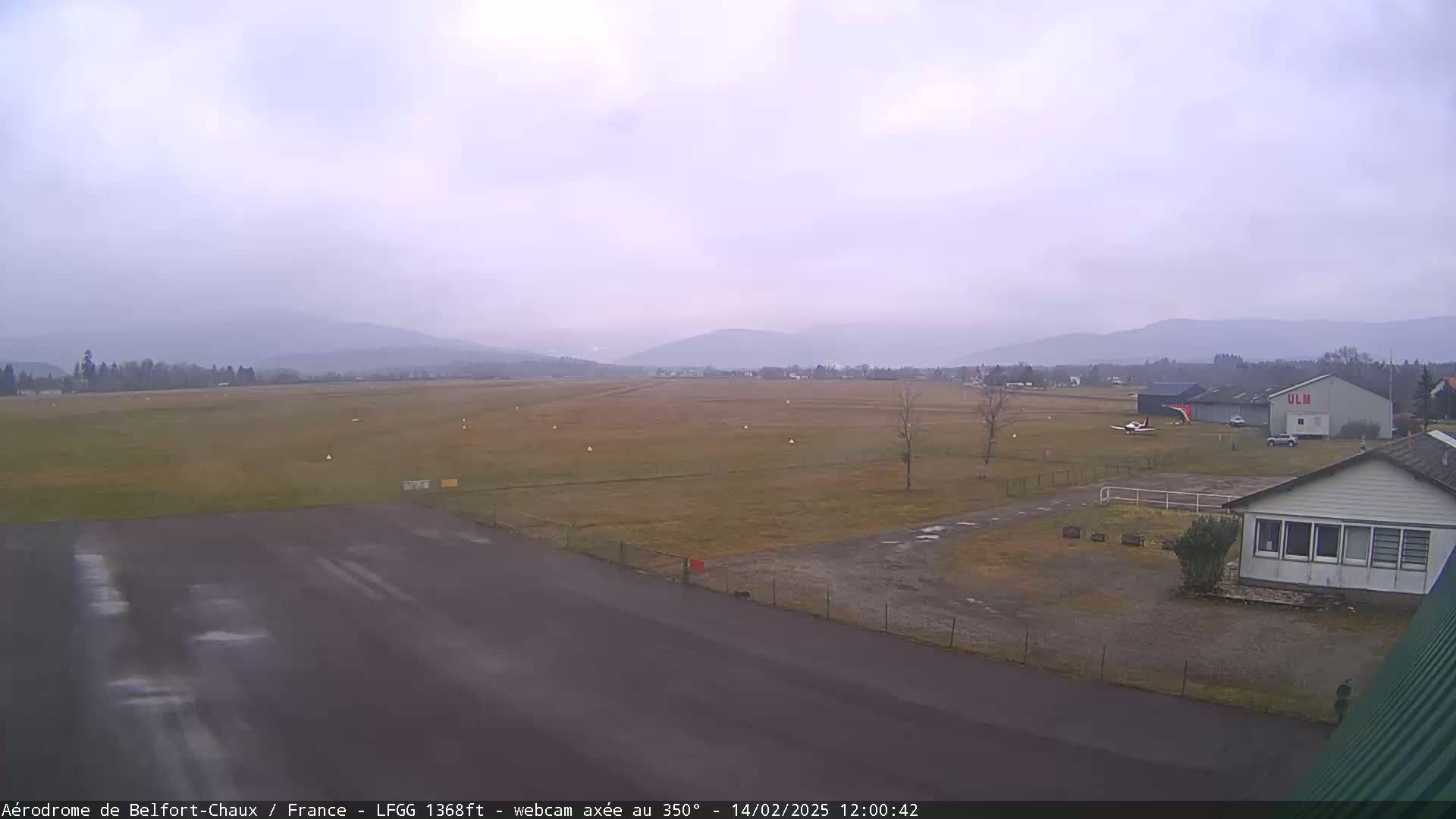 A small airfield with a single-engine airplane and hangar is visible under an overcast sky, with distant, hazy mountains in the background.