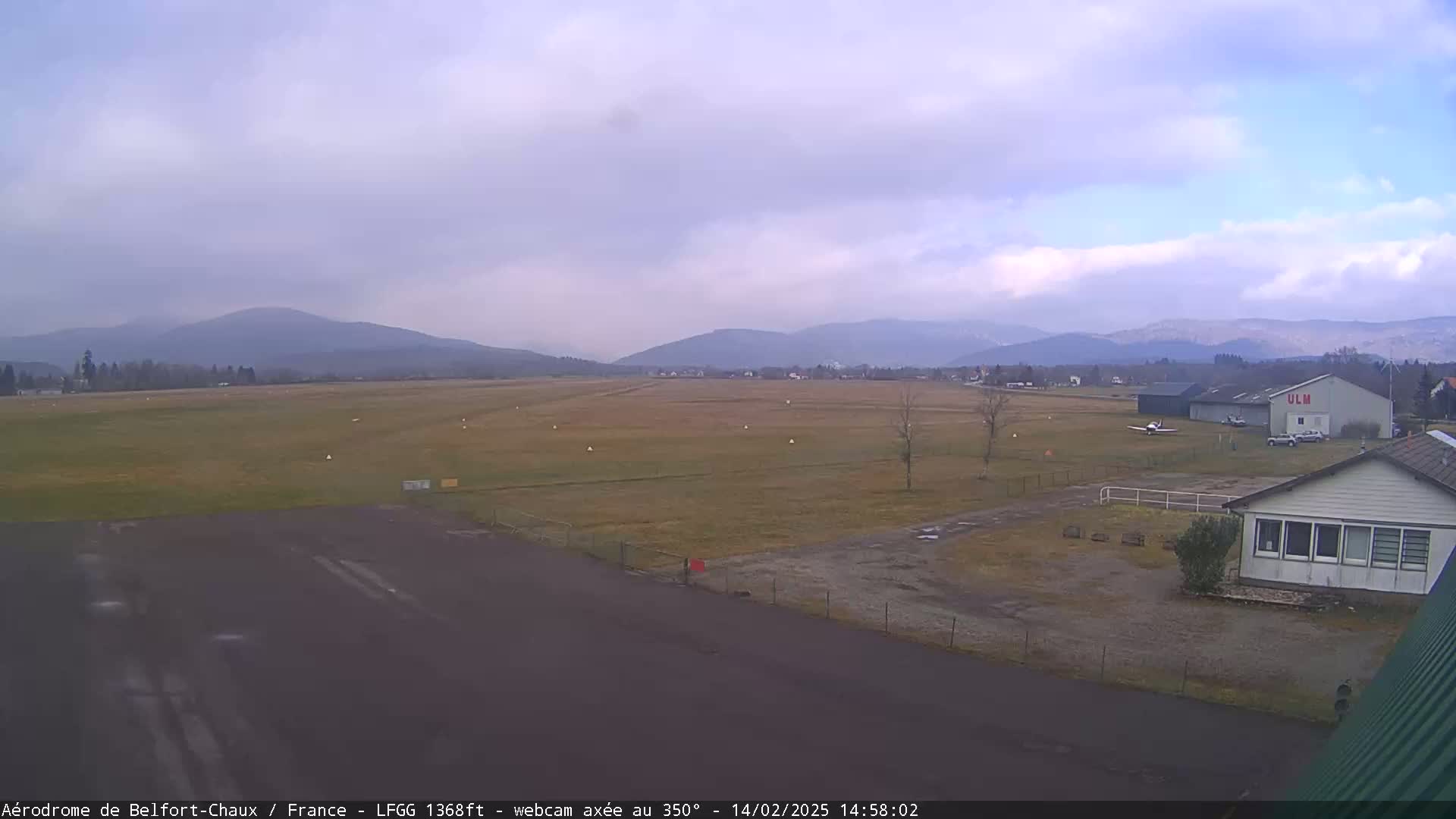 A small airfield with a hangar and a single-story building is situated before a grassy field, mountains in the distance under an overcast sky.
