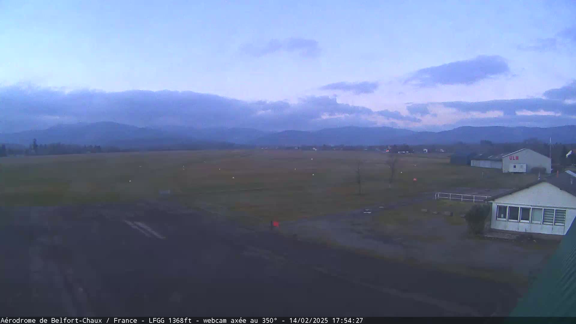 An airfield with a few buildings is shown under a twilight sky with a hazy mountain range in the distance.