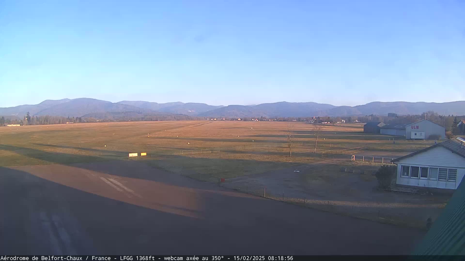 A flat, grassy airfield with a runway and several buildings is visible, under a clear blue sky and bathed in the morning sun, with distant mountains in the background.