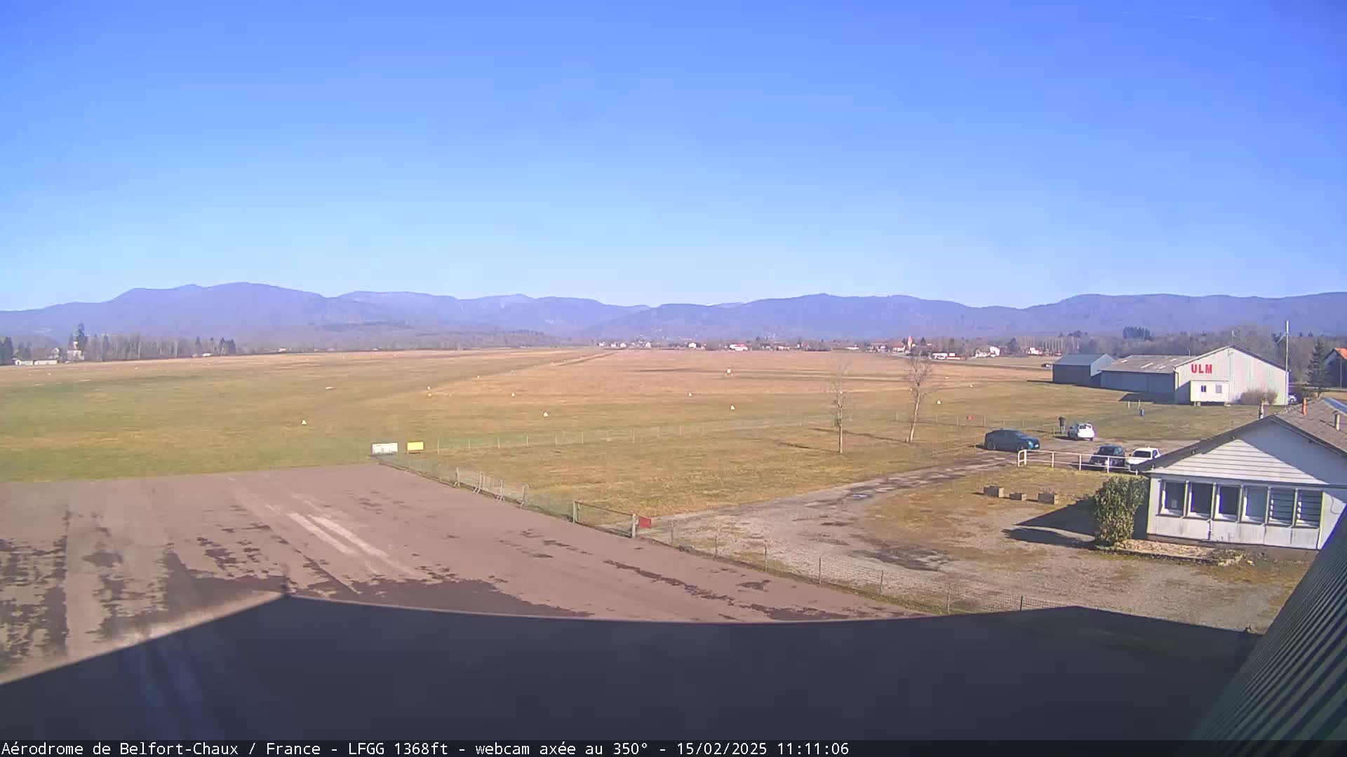A long airfield with a runway and several buildings is visible under a clear blue sky, backed by a range of distant mountains.