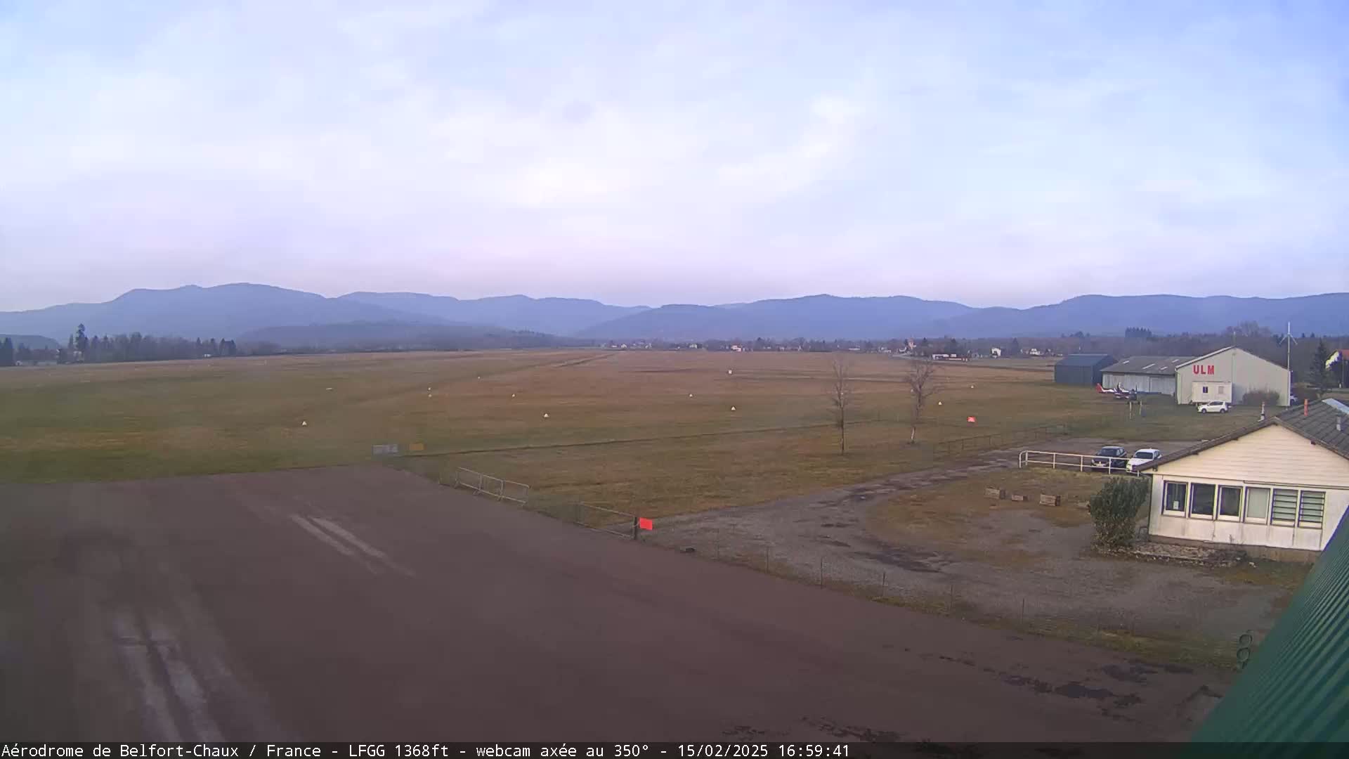 A mostly clear, overcast sky overlooks a flat airfield with a runway and several buildings, backed by distant, hazy mountains.