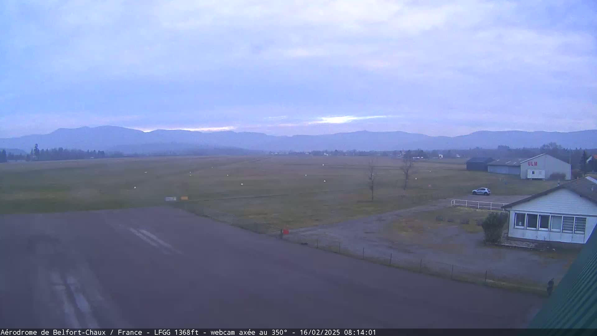 A mostly flat airfield with a few buildings and a car, under a pale, overcast sky with distant mountains visible in the background.