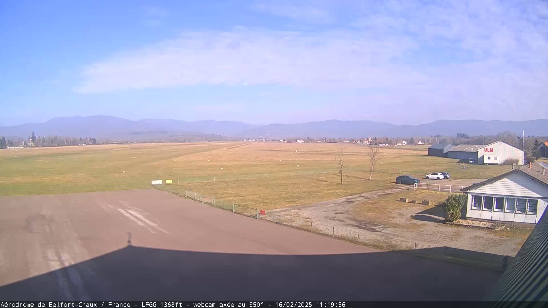 A small airfield with a few cars parked near a building, under a clear blue sky with distant mountains.