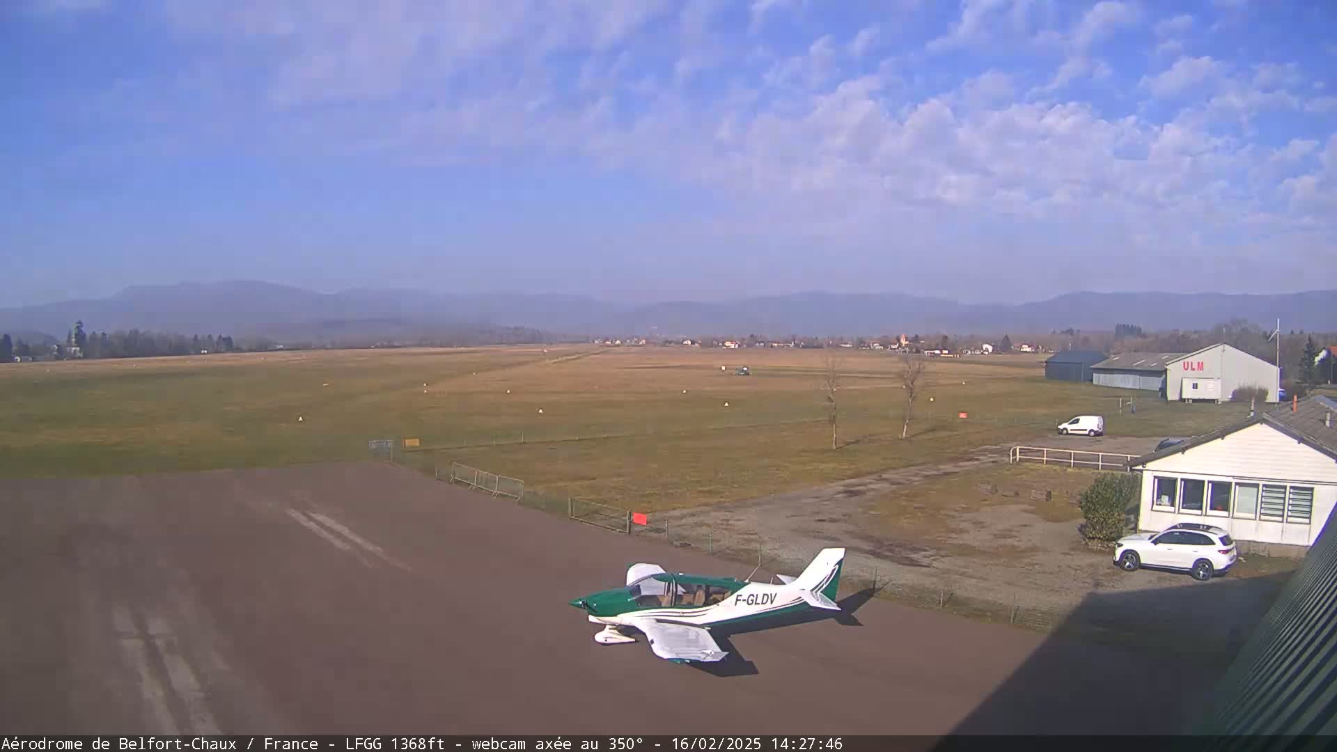 A small airplane sits on a tarmac at a small airport on a partly cloudy day, with distant mountains visible in the background.