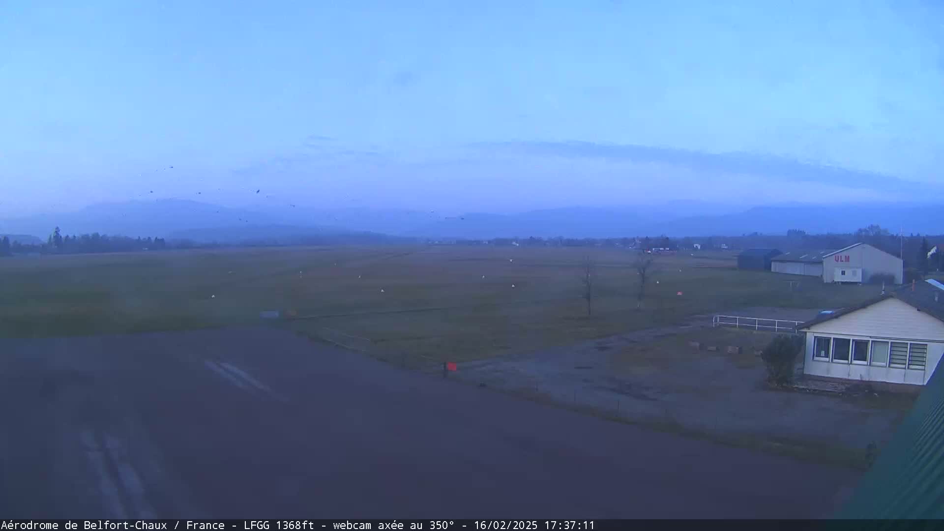 A flat, grassy airfield with a few buildings is visible under a twilight sky with faint clouds and distant mountains.