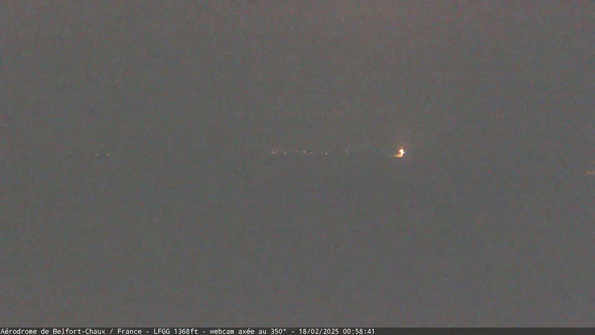 A dark, nighttime scene shows a distant line of faint lights, possibly a town or airport runway, under a cloudy, dark gray sky.