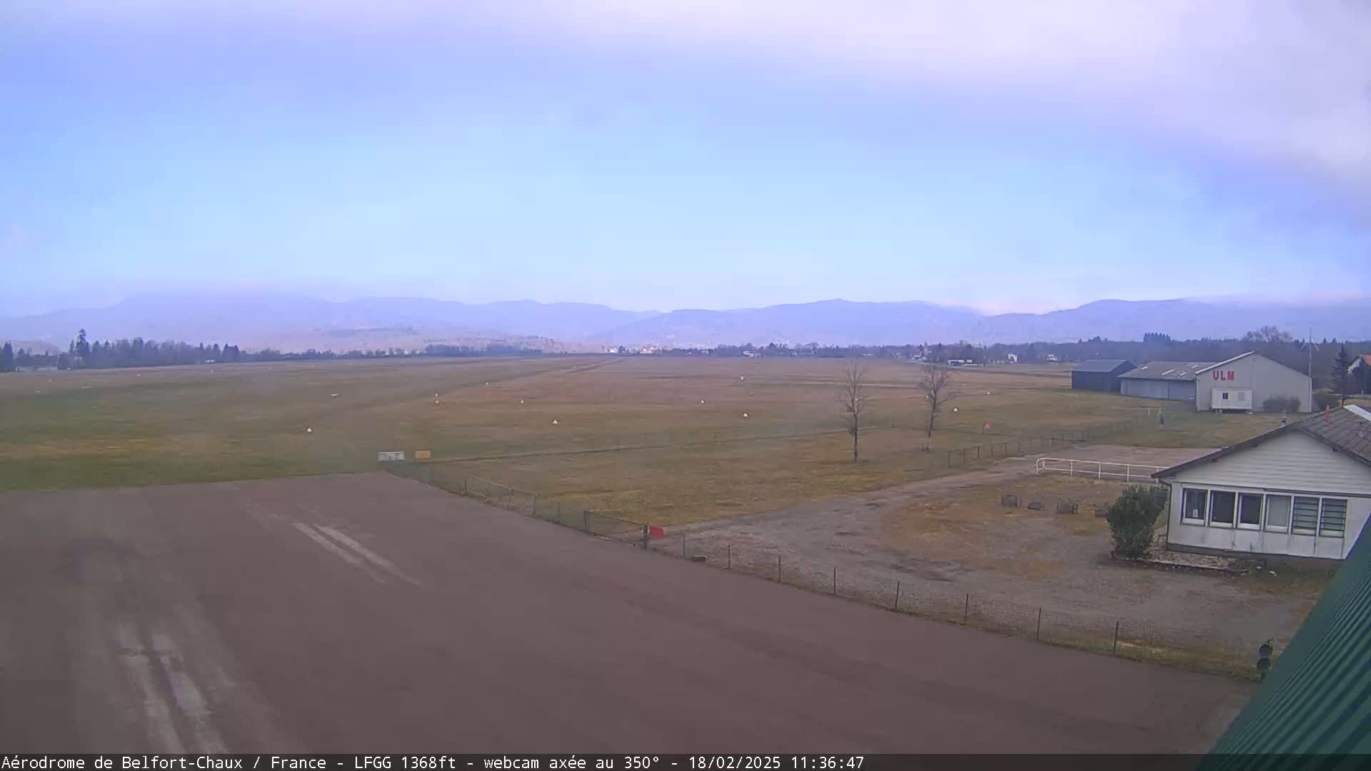 A flat, grassy airfield with a runway and several buildings, is visible under a hazy, overcast sky, with mountains in the background.