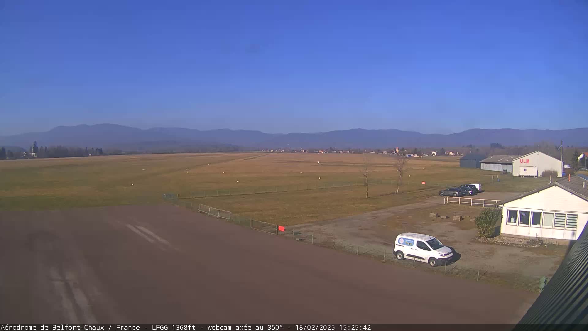 A small airfield with a few vehicles parked near a hangar and some buildings, under a clear blue sky and distant mountains.