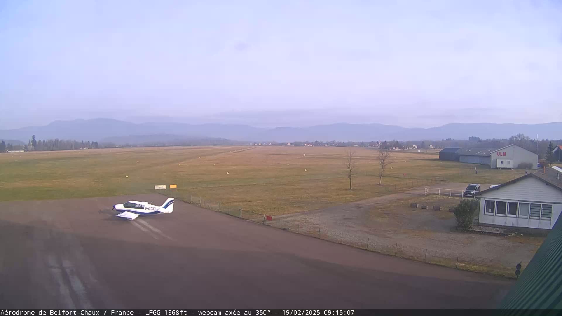 A small airplane sits on a tarmac at an airfield under a clear, light-grey sky, with distant mountains visible in the background.
