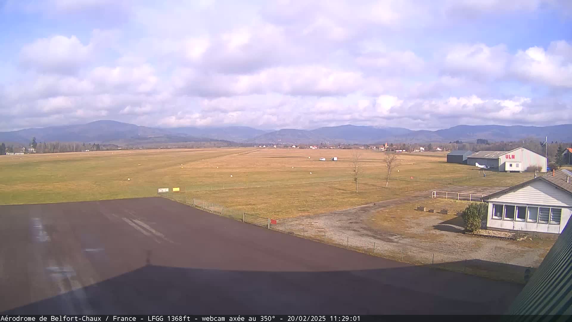 A mostly clear day at a small airfield, with a runway, hangar, and small buildings, and distant mountains in the background.