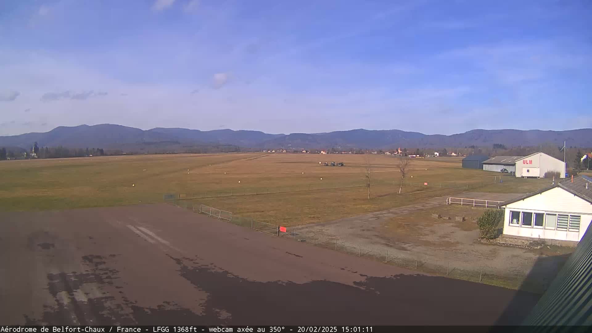 A mostly sunny day at a small airfield shows a runway, several buildings, and distant mountains.
