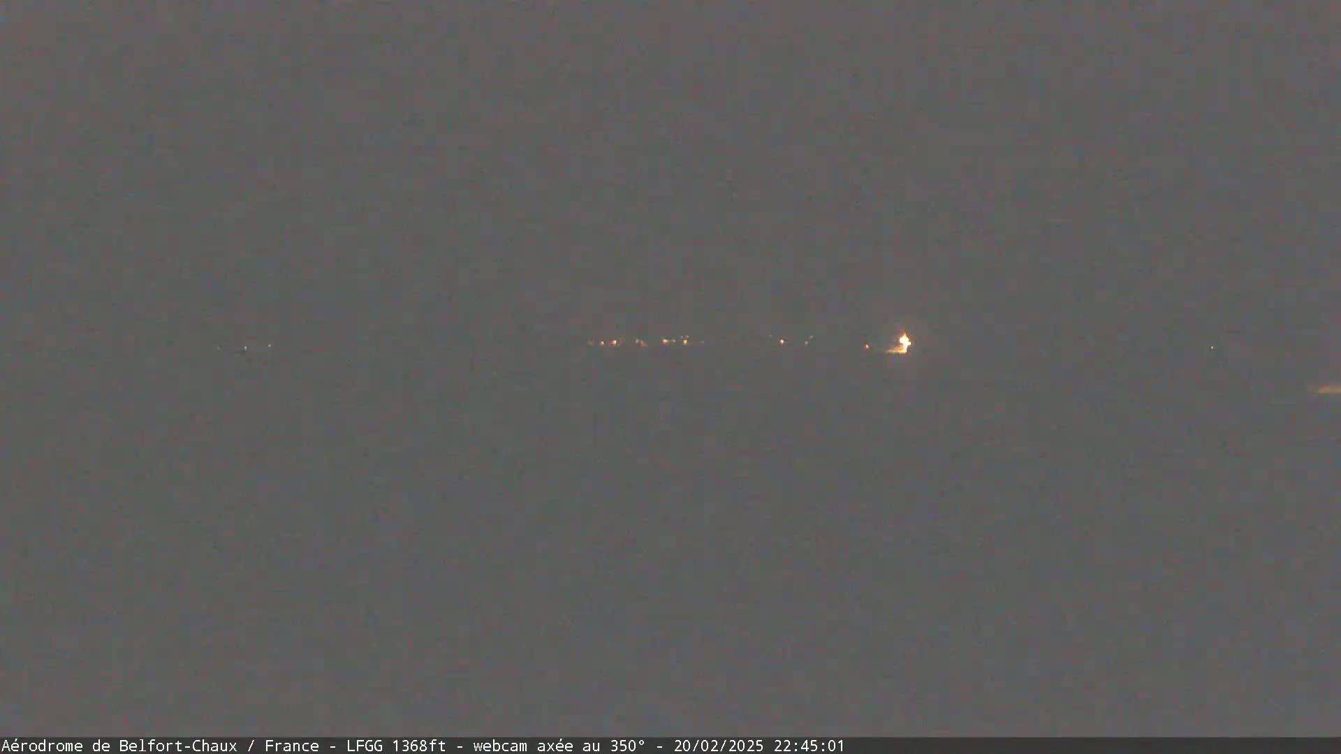 A nighttime long shot shows a dimly lit landscape with a few scattered lights in the distance under a dark, overcast sky.