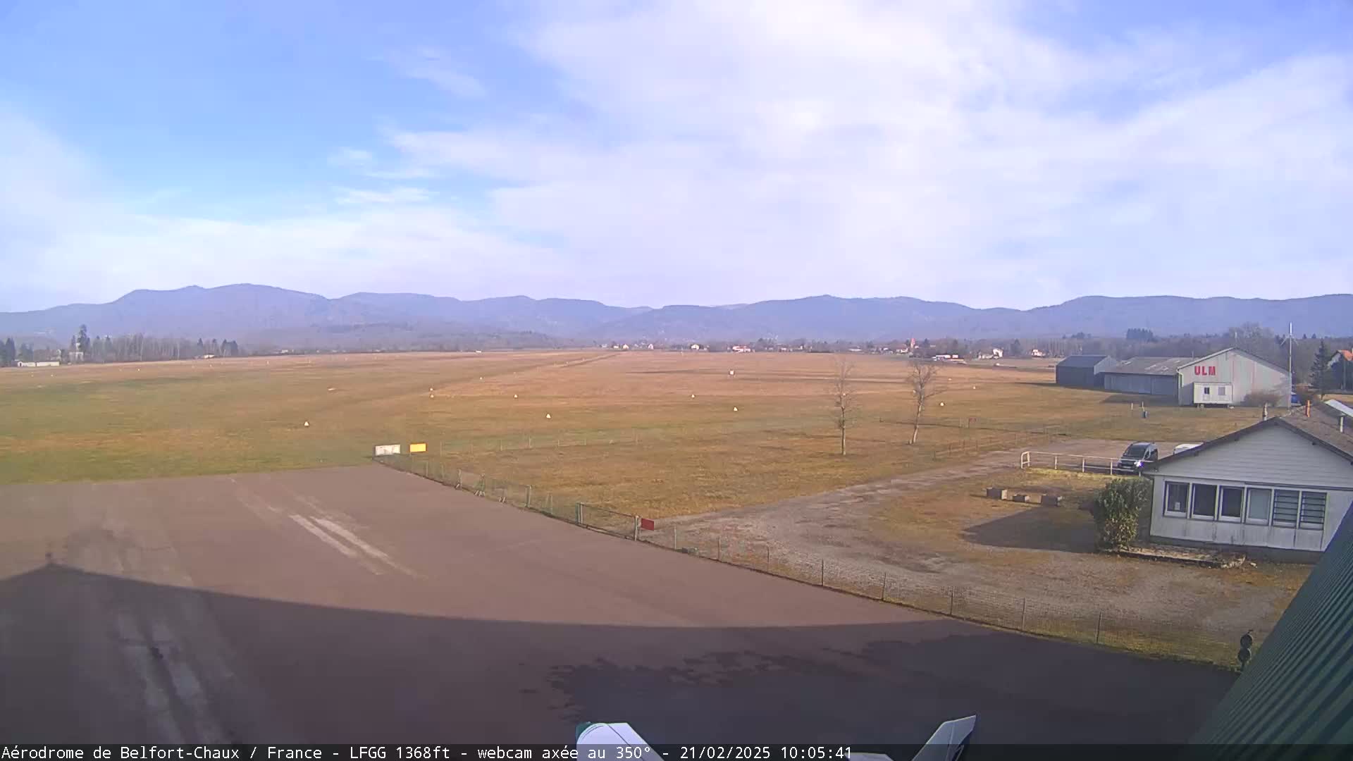 A mostly sunny day at an airfield, with a runway, buildings, and distant mountains.