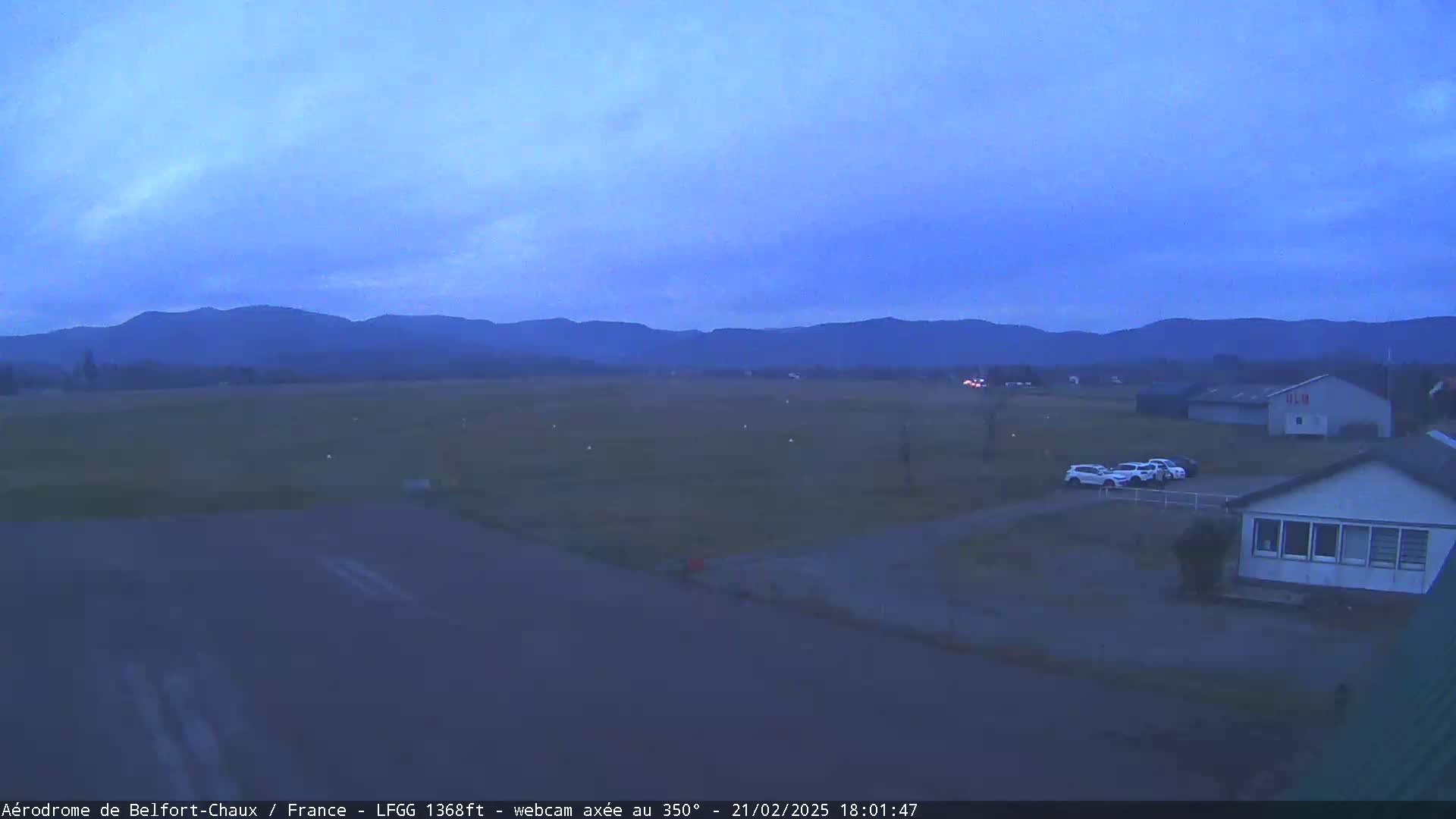 A twilight view of a flat airfield with a few cars parked near a building, mountains visible in the distance under a clear, dark blue sky.