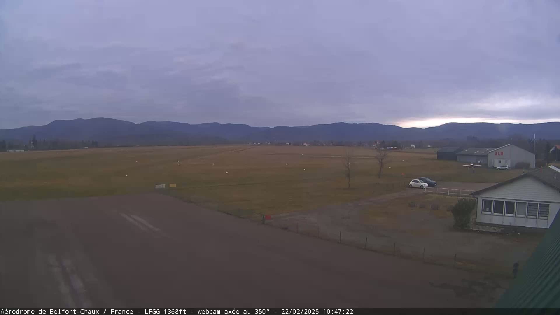 A small airfield with a few cars and a hangar is situated in a flat grassy area, backed by distant mountains under an overcast sky.