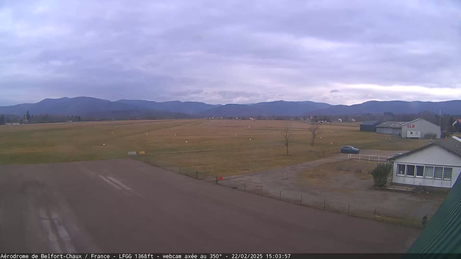 A flat, grassy airfield with a runway and several buildings is visible, under a mostly cloudy sky with distant mountains in the background.