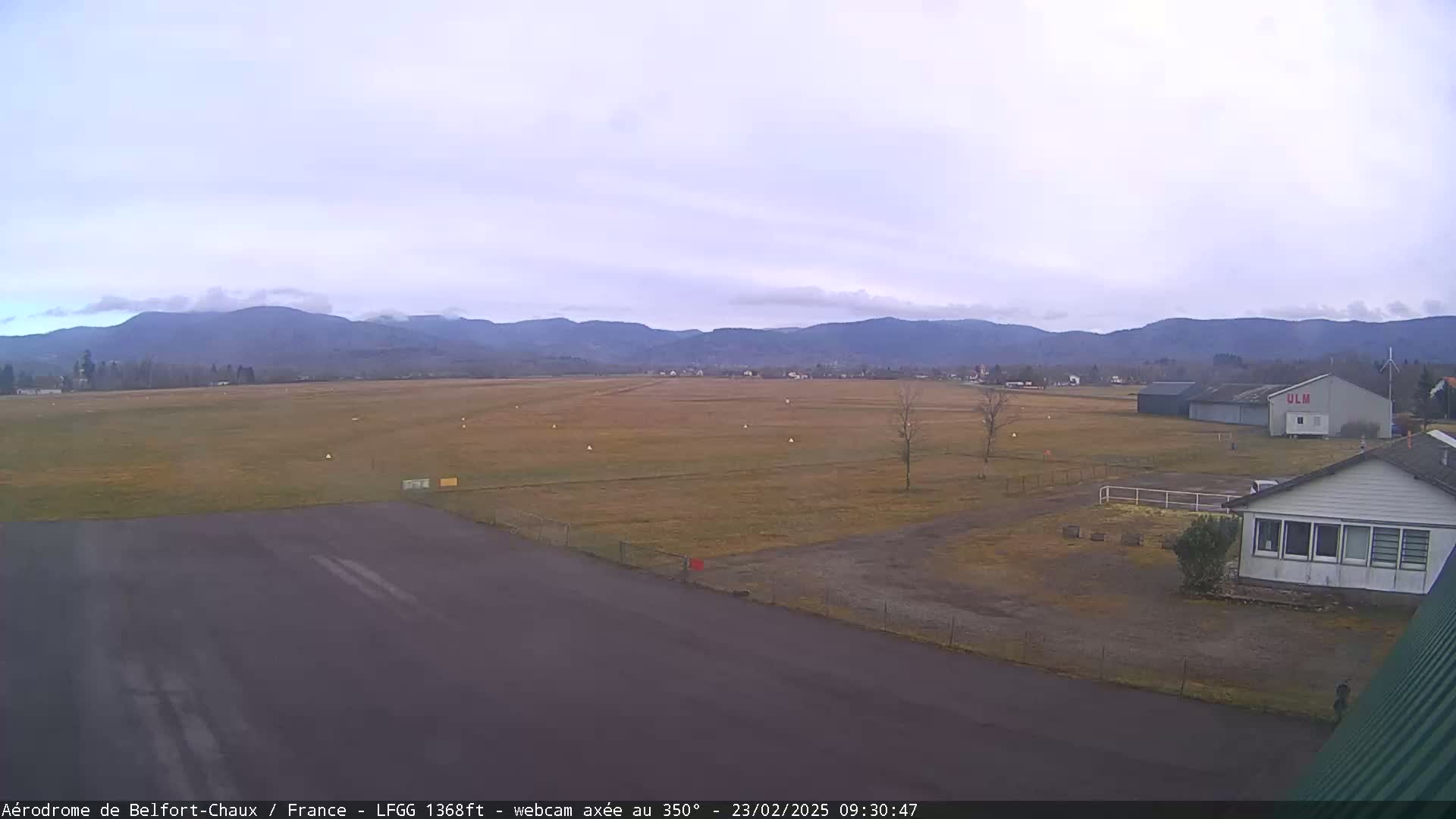 A mostly clear, overcast day reveals a small airfield with buildings and a runway, backed by distant mountains.