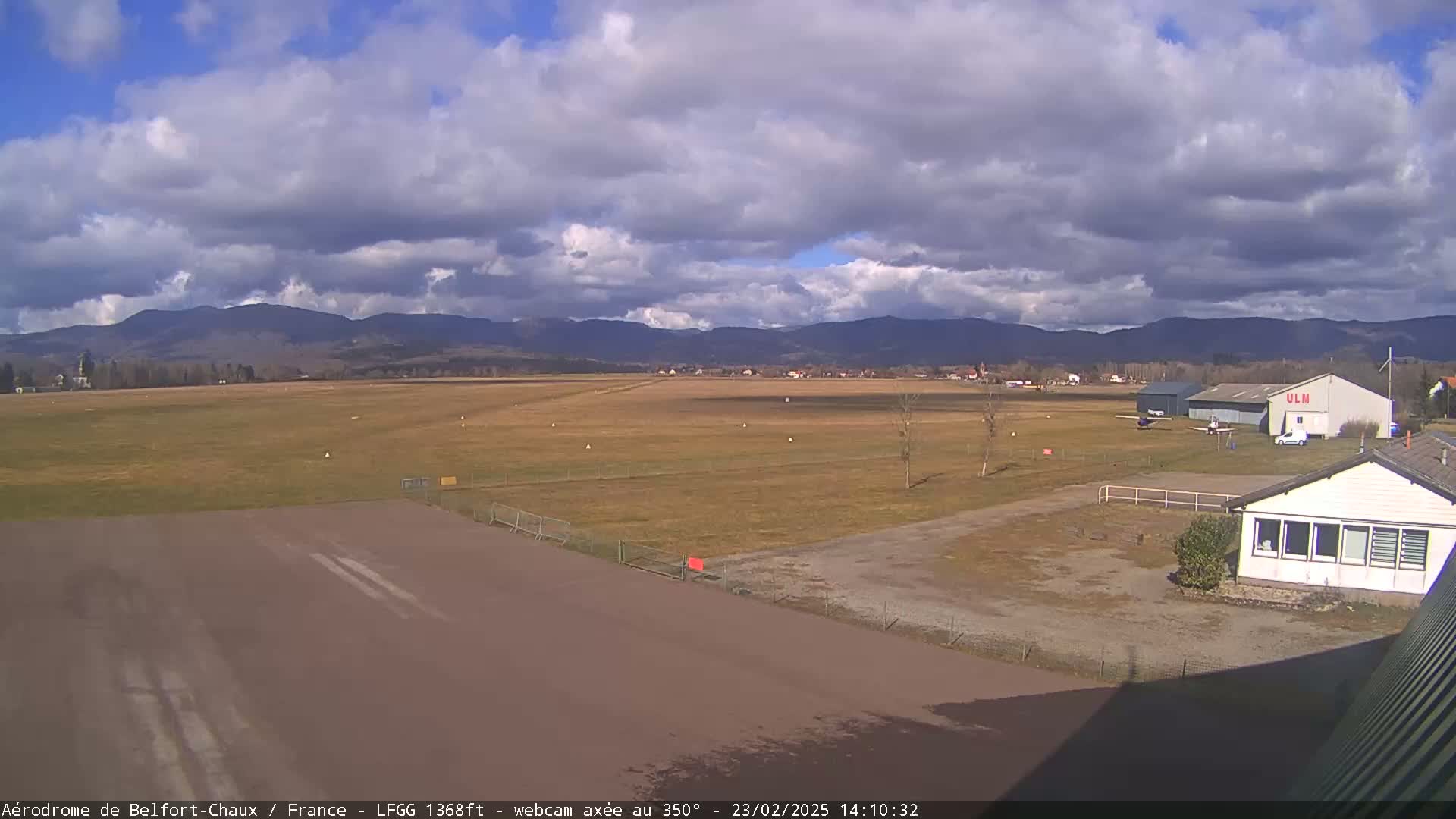 A mostly cloudy day at an airfield shows a runway, small planes, hangars, and distant mountains.