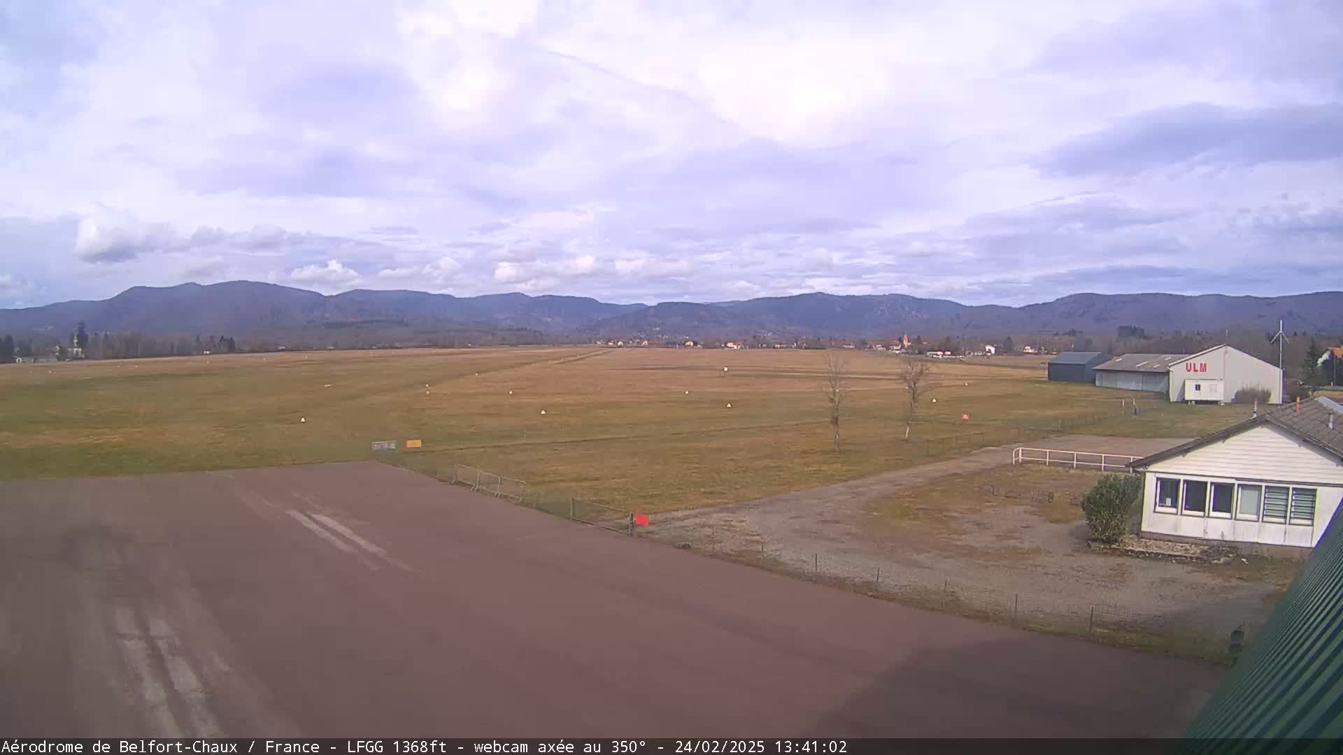 A mostly clear day reveals a flat airfield with a runway, small buildings, and distant mountains under a lightly clouded sky.