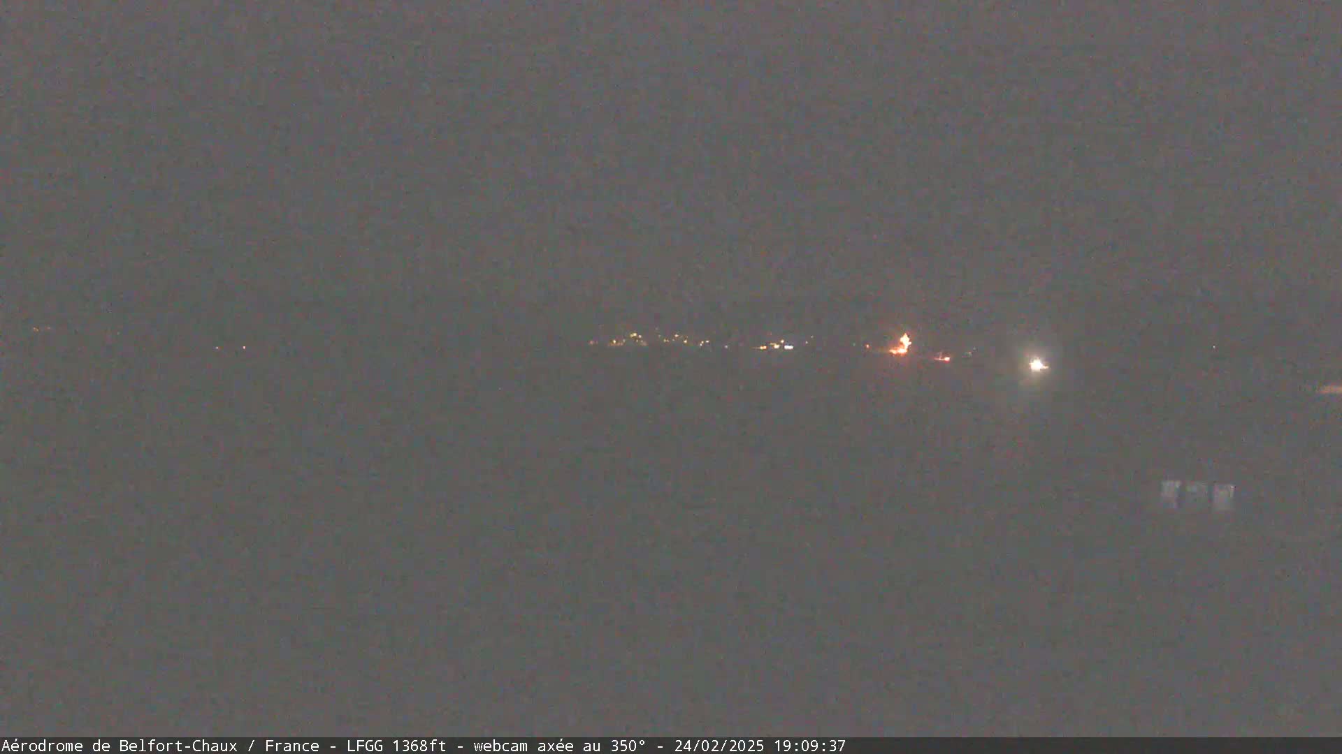 A dark, nighttime scene shows a distant line of faint lights, likely a town or village, under a cloudy sky.
